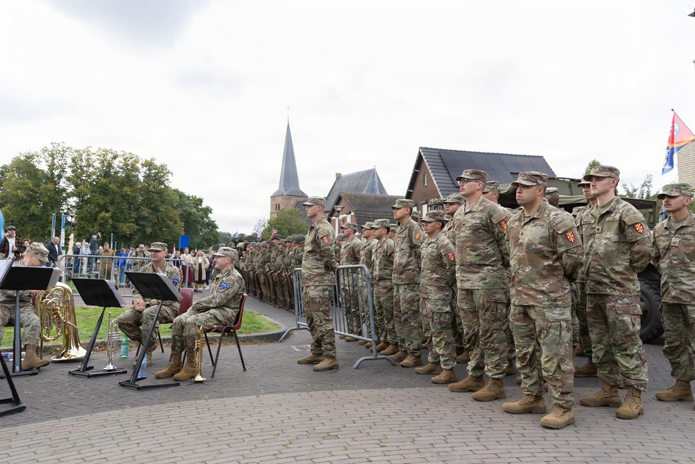A formation of military personnel in camouflage uniforms stands at attention during what appears to be a formal outdoor ceremony in a European town setting. A military band with brass instruments is visible on the left side, with seated officers nearby. Civilians and spectators are gathered in the background near metal crowd-control barriers.