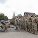 A formation of military personnel in camouflage uniforms stands at attention during what appears to be a formal outdoor ceremony in a European town setting. A military band with brass instruments is visible on the left side, with seated officers nearby. Civilians and spectators are gathered in the background near metal crowd-control barriers.