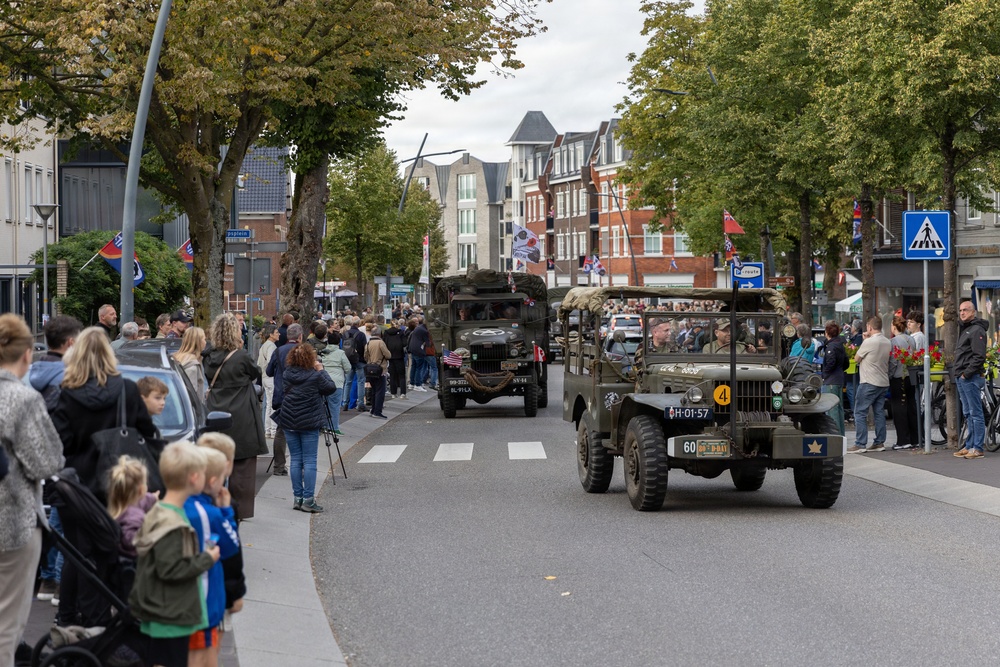 A commemorative military parade is taking place on a tree-lined urban street, featuring vintage WWII-era American military vehicles including a Willys Jeep and a military truck. Crowds of spectators line both sides of the street to watch the procession. The Dutch license plates and street signage suggest this event is taking place in the Netherlands, likely a liberation commemoration.