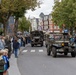 A commemorative military parade is taking place on a tree-lined urban street, featuring vintage WWII-era American military vehicles including a Willys Jeep and a military truck. Crowds of spectators line both sides of the street to watch the procession. The Dutch license plates and street signage suggest this event is taking place in the Netherlands, likely a liberation commemoration.