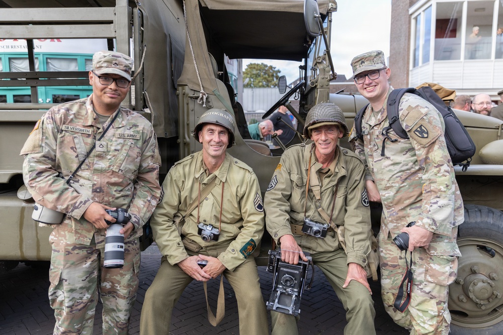 Four individuals pose in front of a vintage military truck, two wearing modern U.S. Army MultiCam (OCP) uniforms and two dressed in WWII-era U.S. Army photographer uniforms with steel helmets marked 'PHOTOGRAPHER'. All four are holding cameras, suggesting a photography-themed military commemoration or historical reenactment event. The name tape 'SILVERSMITH' is visible on the leftmost modern soldier.