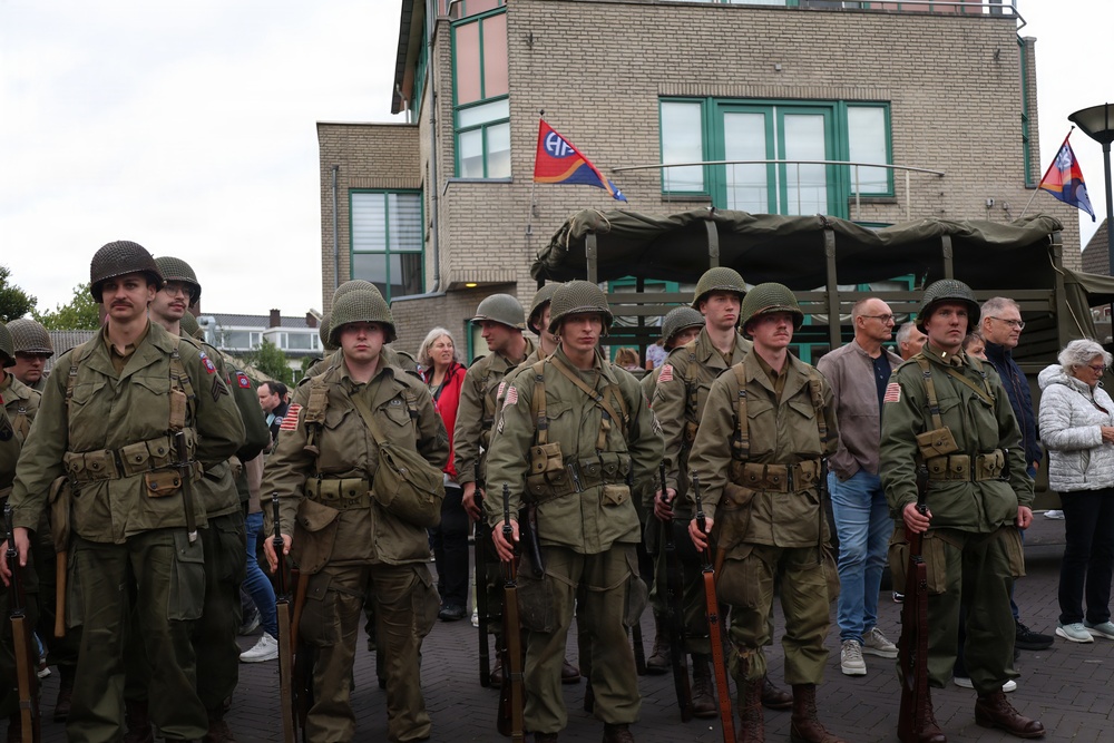 A group of individuals dressed in WWII-era U.S. Army uniforms are participating in what appears to be a historical reenactment or commemorative event. They are wearing M1 steel helmets, olive drab uniforms with American flag patches, and carrying period-correct rifles. Civilian spectators and a military truck with a canvas cover are visible in the background, along with flags including what appears to be an 82nd Airborne Division pennant.
