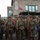 A group of individuals dressed in WWII-era U.S. Army uniforms are participating in what appears to be a historical reenactment or commemorative event. They are wearing M1 steel helmets, olive drab uniforms with American flag patches, and carrying period-correct rifles. Civilian spectators and a military truck with a canvas cover are visible in the background, along with flags including what appears to be an 82nd Airborne Division pennant.