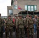 A group of individuals dressed in WWII-era U.S. Army uniforms are participating in what appears to be a historical reenactment or commemorative event. They are wearing M1 steel helmets, olive drab uniforms with American flag patches, and carrying period-correct rifles. Civilian spectators and a military truck with a canvas cover are visible in the background, along with flags including what appears to be an 82nd Airborne Division pennant.