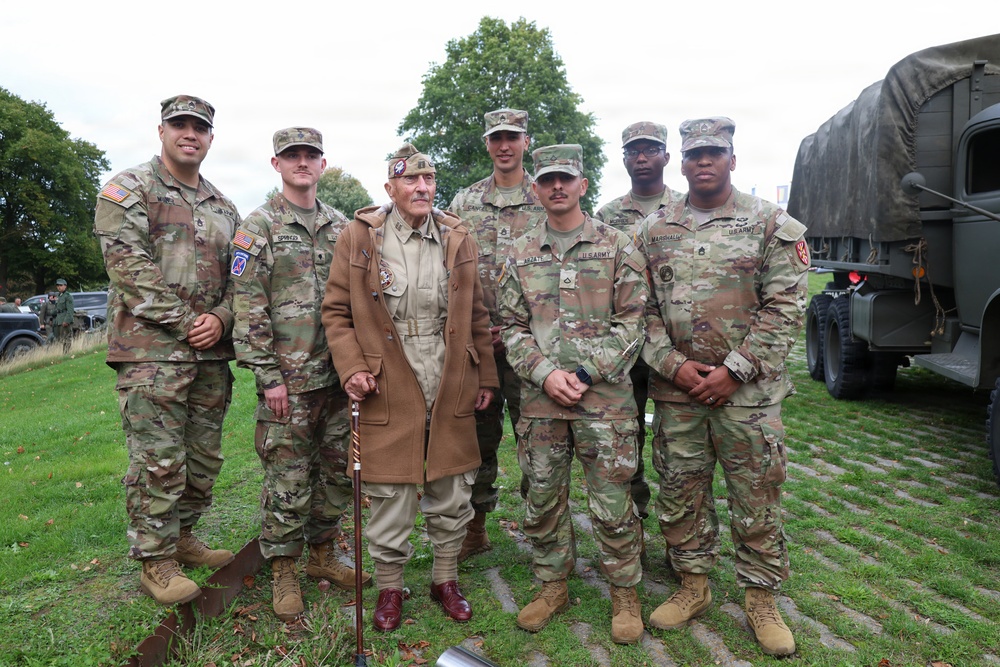 A group of six active-duty U.S. Army soldiers in OCP (Operational Camouflage Pattern) uniforms pose with an elderly veteran dressed in WWII-era attire, including a tan uniform and garrison cap with insignia. The group is gathered outdoors on a grassy area near a military vehicle, likely at a commemorative or memorial event. The elderly individual appears to be a honored guest or veteran being recognized at the gathering.