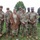 A group of six active-duty U.S. Army soldiers in OCP (Operational Camouflage Pattern) uniforms pose with an elderly veteran dressed in WWII-era attire, including a tan uniform and garrison cap with insignia. The group is gathered outdoors on a grassy area near a military vehicle, likely at a commemorative or memorial event. The elderly individual appears to be a honored guest or veteran being recognized at the gathering.