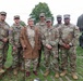 A group of six active-duty U.S. Army soldiers in OCP (Operational Camouflage Pattern) uniforms pose with an elderly veteran dressed in WWII-era attire, including a tan uniform and garrison cap with insignia. The group is gathered outdoors on a grassy area near a military vehicle, likely at a commemorative or memorial event. The elderly individual appears to be a honored guest or veteran being recognized at the gathering.