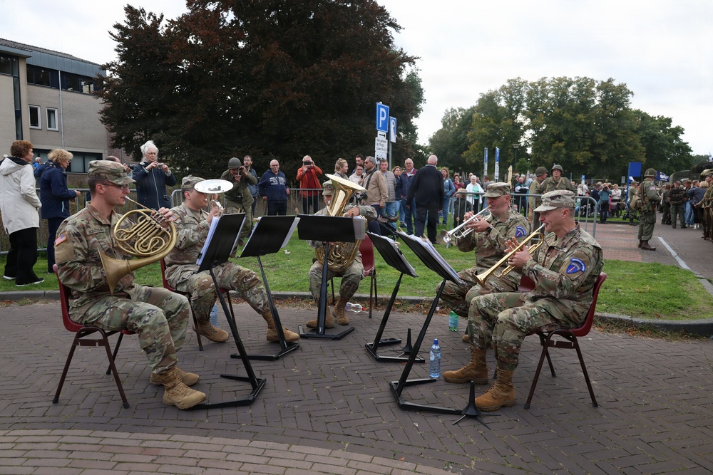 A military brass ensemble of approximately five soldiers in camouflage uniforms performs outdoors on a brick plaza for a crowd of civilian spectators. The musicians are seated in a semicircle with music stands, playing instruments including a French horn, trumpet, and euphonium/tuba. The setting appears to be a public commemorative or ceremonial event, possibly in the Netherlands based on visible Dutch parking signage.