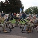 A military brass ensemble of approximately five soldiers in camouflage uniforms performs outdoors on a brick plaza for a crowd of civilian spectators. The musicians are seated in a semicircle with music stands, playing instruments including a French horn, trumpet, and euphonium/tuba. The setting appears to be a public commemorative or ceremonial event, possibly in the Netherlands based on visible Dutch parking signage.
