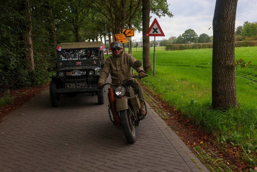 A vintage military motorcycle and a WWII-era Willys Jeep travel along a brick-paved road lined with trees in what appears to be the Netherlands. The rider is dressed in period-appropriate military-style clothing with goggles and a helmet. The scene likely depicts a historical vehicle rally or commemoration event.