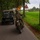 A vintage military motorcycle and a WWII-era Willys Jeep travel along a brick-paved road lined with trees in what appears to be the Netherlands. The rider is dressed in period-appropriate military-style clothing with goggles and a helmet. The scene likely depicts a historical vehicle rally or commemoration event.