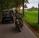 A vintage military motorcycle and a WWII-era Willys Jeep travel along a brick-paved road lined with trees in what appears to be the Netherlands. The rider is dressed in period-appropriate military-style clothing with goggles and a helmet. The scene likely depicts a historical vehicle rally or commemoration event.