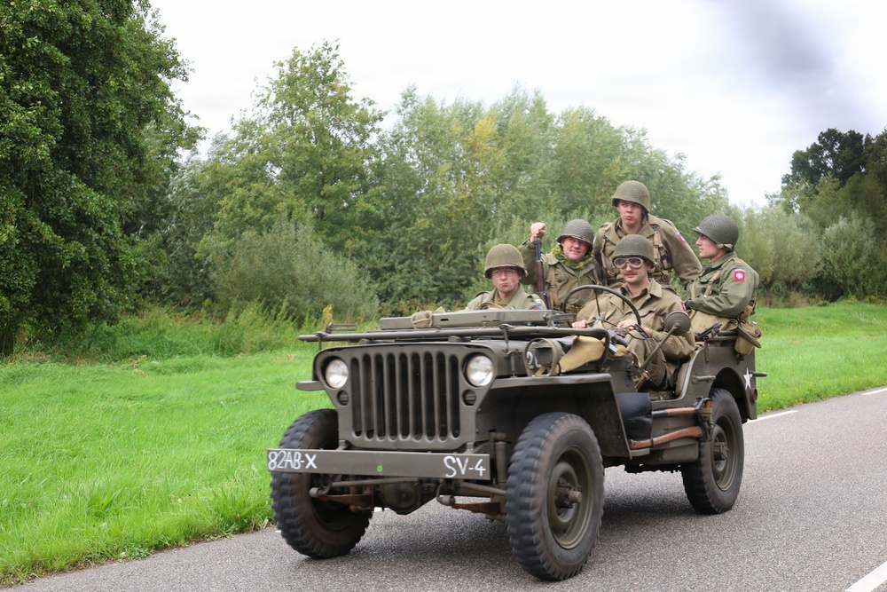 A group of individuals dressed in WWII-era U.S. Army uniforms and helmets are riding in an olive-drab Willys MB military jeep on a rural road. The vehicle displays markings '82AB-X' and 'SV-4', consistent with 82nd Airborne Division reenactment insignia. The setting and equipment suggest this is a historical reenactment or commemorative event.
