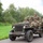 A group of individuals dressed in WWII-era U.S. Army uniforms and helmets are riding in an olive-drab Willys MB military jeep on a rural road. The vehicle displays markings '82AB-X' and 'SV-4', consistent with 82nd Airborne Division reenactment insignia. The setting and equipment suggest this is a historical reenactment or commemorative event.