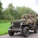 A group of individuals dressed in WWII-era U.S. Army uniforms and helmets are riding in an olive-drab Willys MB military jeep on a rural road. The vehicle displays markings '82AB-X' and 'SV-4', consistent with 82nd Airborne Division reenactment insignia. The setting and equipment suggest this is a historical reenactment or commemorative event.