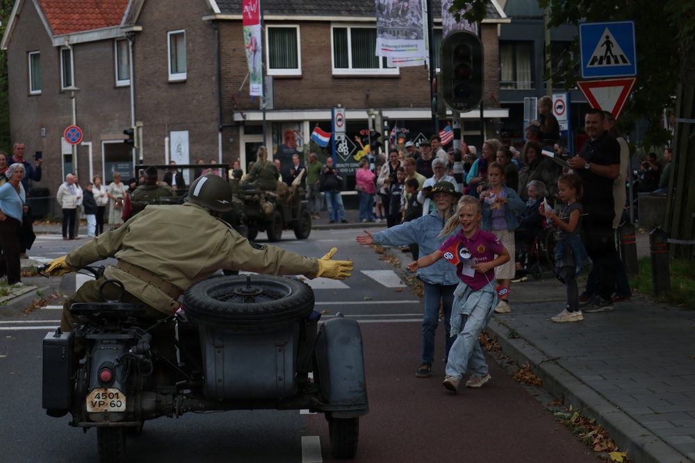 A historical military parade or commemoration event taking place on a Dutch street, featuring a person dressed in WWII-era Allied military uniform riding a vintage motorcycle with sidecar. The rider is reaching out toward excited children on the sidewalk, while a large crowd of spectators lines the street. Dutch and American flags are visible in the background, suggesting a liberation commemoration event.