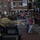A historical military parade or commemoration event taking place on a Dutch street, featuring a person dressed in WWII-era Allied military uniform riding a vintage motorcycle with sidecar. The rider is reaching out toward excited children on the sidewalk, while a large crowd of spectators lines the street. Dutch and American flags are visible in the background, suggesting a liberation commemoration event.