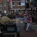 A historical military parade or commemoration event taking place on a Dutch street, featuring a person dressed in WWII-era Allied military uniform riding a vintage motorcycle with sidecar. The rider is reaching out toward excited children on the sidewalk, while a large crowd of spectators lines the street. Dutch and American flags are visible in the background, suggesting a liberation commemoration event.