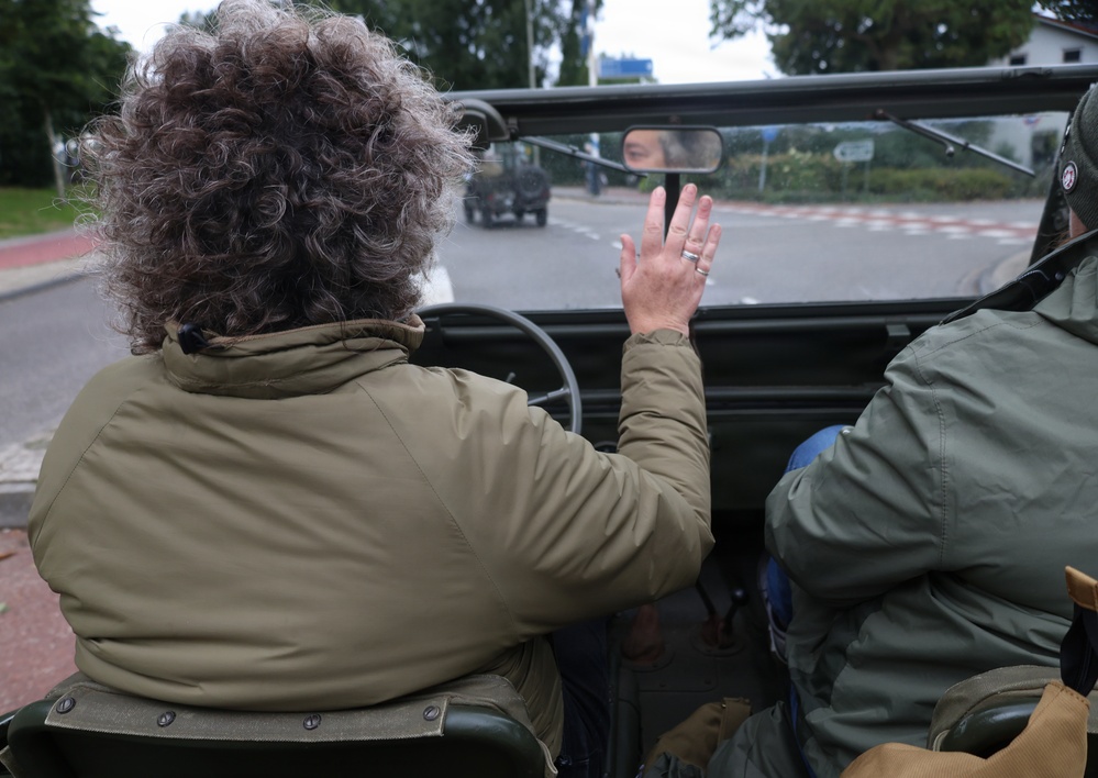 A person with curly gray-brown hair, wearing an olive/khaki jacket, is seated in an open-top military-style vehicle, raising their hand in a waving gesture. Another person in a similar green jacket is seated to the right. The vehicle is traveling on a road, following another military-style vehicle visible ahead.