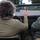 A person with curly gray-brown hair, wearing an olive/khaki jacket, is seated in an open-top military-style vehicle, raising their hand in a waving gesture. Another person in a similar green jacket is seated to the right. The vehicle is traveling on a road, following another military-style vehicle visible ahead.