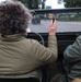 A person with curly gray-brown hair, wearing an olive/khaki jacket, is seated in an open-top military-style vehicle, raising their hand in a waving gesture. Another person in a similar green jacket is seated to the right. The vehicle is traveling on a road, following another military-style vehicle visible ahead.