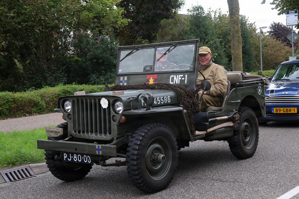 An elderly man drives a restored WWII-era Willys MB military jeep on a public road, likely during a historical vehicle parade or commemorative event. The olive-drab jeep bears military markings, unit insignia, and camouflage netting, along with a Dutch license plate 'PJ-80-00'. A blue Mini Cooper is visible parked in the background.