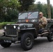 An elderly man drives a restored WWII-era Willys MB military jeep on a public road, likely during a historical vehicle parade or commemorative event. The olive-drab jeep bears military markings, unit insignia, and camouflage netting, along with a Dutch license plate 'PJ-80-00'. A blue Mini Cooper is visible parked in the background.