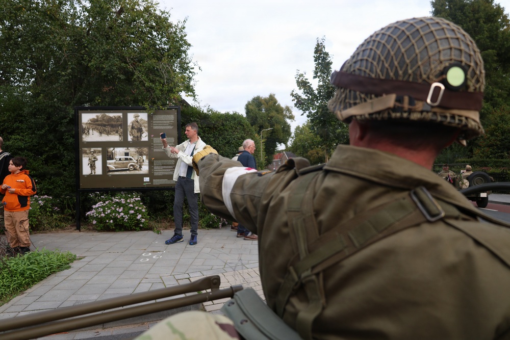 The image depicts what appears to be a World War II commemorative or reenactment event in a public outdoor space. A person in period-accurate WWII Allied military uniform and helmet is visible in the foreground, while civilians gather around an outdoor historical display board featuring archival photographs. The setting appears to be a European town square or memorial area.