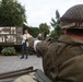 The image depicts what appears to be a World War II commemorative or reenactment event in a public outdoor space. A person in period-accurate WWII Allied military uniform and helmet is visible in the foreground, while civilians gather around an outdoor historical display board featuring archival photographs. The setting appears to be a European town square or memorial area.