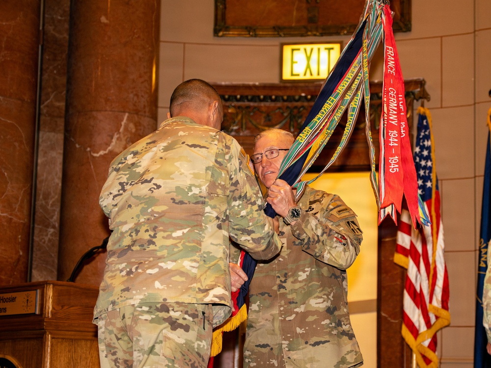 Two U.S. Army soldiers in OCP (Operational Camouflage Pattern) uniforms are handling a military unit guidon or colors with multiple campaign streamers attached. The ceremony appears to be taking place in a formal indoor venue, likely a state capitol or government building, with an American flag and what appears to be a state flag visible in the background. Campaign streamers visible include one reading 'France-Germany 1944-1945', indicating WWII-era battle honors.