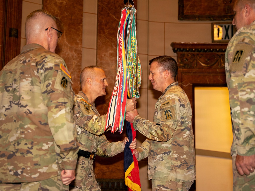 A military change of command ceremony is taking place indoors, with two soldiers in U.S. Army OCP (Operational Camouflage Pattern) uniforms exchanging a unit guidon (flag) adorned with multiple campaign streamers. The ceremony appears formal, conducted in an ornate interior space. Additional soldiers are present as witnesses to the transfer of command.