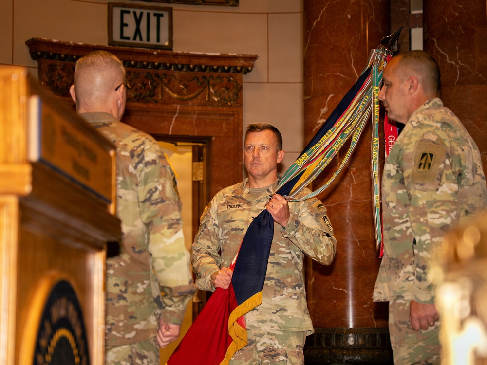 Three U.S. Army soldiers in OCP (Operational Camouflage Pattern) uniforms are participating in a formal military ceremony, likely a change of command or activation/inactivation ceremony. The central figure, identified by nametape as 'TOOLEY,' is holding an unfurled guidon or organizational flag along with multiple campaign or unit streamers. The event is taking place in an ornate indoor venue with marble columns and decorative woodwork.