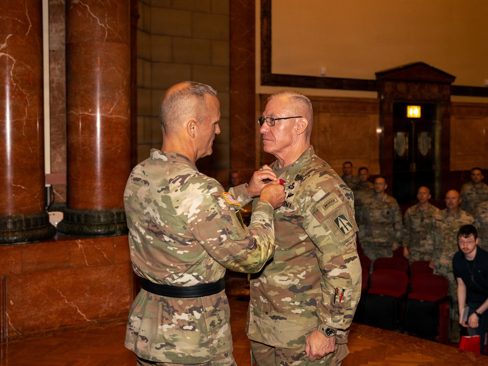 A military award ceremony is taking place in a formal indoor venue with marble columns. One soldier is pinning a medal onto the chest of another soldier while an audience of uniformed personnel observes from the background. Both individuals are wearing U.S. Army OCP (Operational Camouflage Pattern) uniforms.