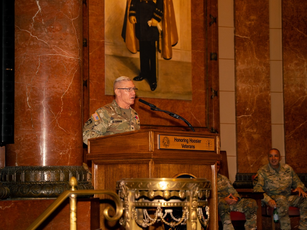 A military officer identified by nameplate as 'EAKEN' is delivering a speech at a wooden podium bearing the text 'Honoring Hoosier Veterans' during what appears to be a veterans recognition ceremony. The setting is an ornate marble-columned hall, likely the Indiana State Capitol or a similar government building. A second military personnel member is seated in the background to the right.