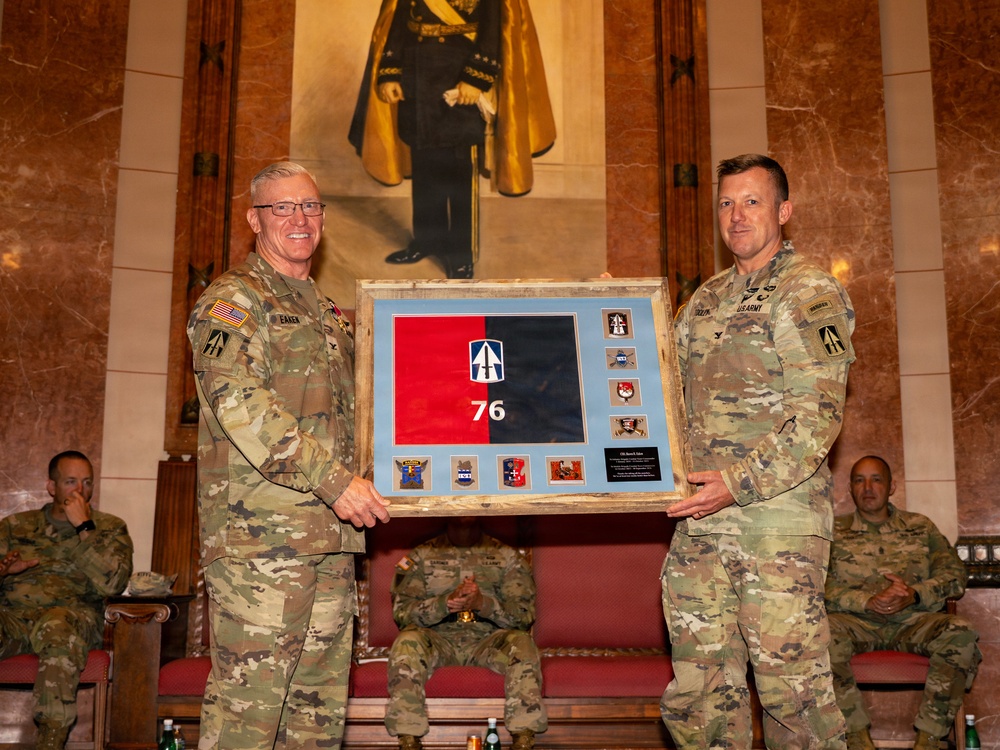 Two U.S. Army soldiers in OCP (Operational Camouflage Pattern) uniforms are presenting a framed shadow box containing a military flag and unit patches. The framed display features a red and black flag with the number '76' and multiple unit insignia patches arranged around it. The ceremony appears to be taking place in an ornate hall with marble walls and a large portrait painting in the background.