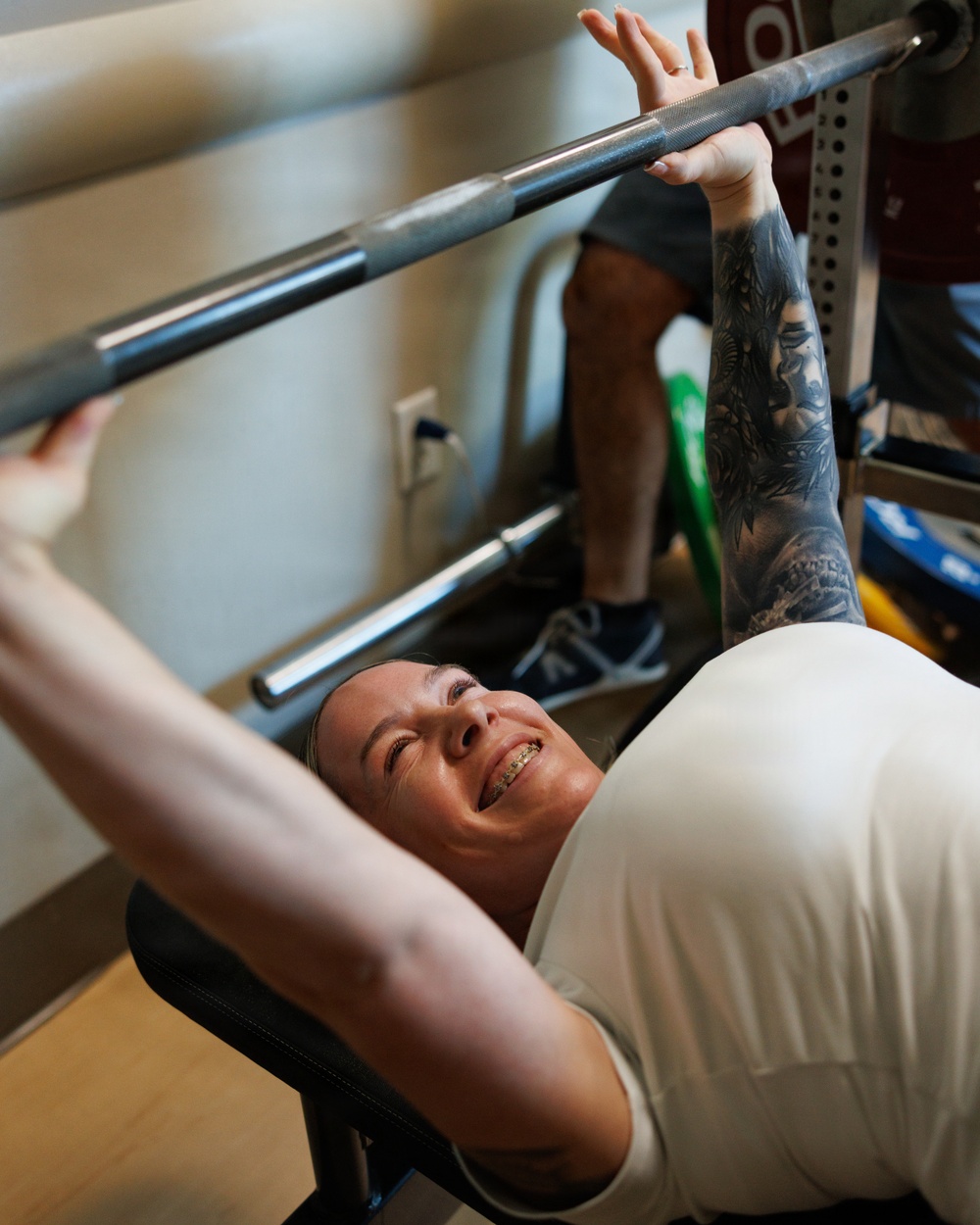 A woman is performing a bench press exercise in what appears to be a home or small gym setting. She is lying on a bench, gripping a barbell with both arms extended, and has extensive tattoos covering her right arm. A second person is visible standing in the background, likely acting as a spotter.