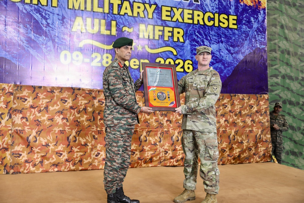 Two military officers, one in Indian Army uniform and one in U.S. Army uniform, are jointly holding a commemorative plaque or award during a ceremony. The event is identified as a Joint Military Exercise named 'AULI - MFFR' scheduled from 09-28 of a month in 2026. The ceremony appears to be a formal exchange or presentation marking the exercise.