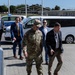 A group of individuals, including a U.S. Army soldier in multicam uniform and several civilians in business attire, are walking across a paved area at what appears to be a military installation. A Military Police vehicle is visible in the background along with a white passenger van. The setting suggests an official visit or delegation tour at a military base.