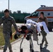 Two military personnel in camouflage uniforms are transporting a patient on a wheeled stretcher across a paved area. In the background, a Romanian ambulance ('Ambulanța') with orange and white markings is visible. This appears to be a military medical exercise or real casualty evacuation scenario.