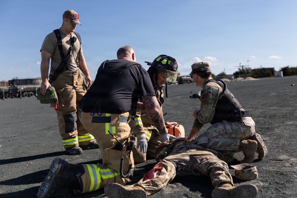 A joint emergency response training exercise is taking place on an open tarmac or gravel surface. Firefighters in turnout gear and military personnel in camouflage uniforms are practicing casualty care on a simulated victim lying on the ground. The scene depicts coordinated first responder and military medical response procedures.