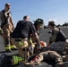 A joint emergency response training exercise is taking place on an open tarmac or gravel surface. Firefighters in turnout gear and military personnel in camouflage uniforms are practicing casualty care on a simulated victim lying on the ground. The scene depicts coordinated first responder and military medical response procedures.