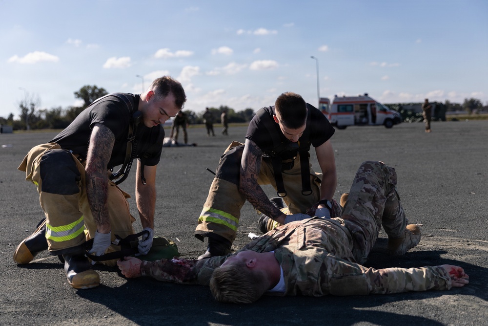 Two individuals in firefighter turnout gear (pants with reflective yellow stripes) and black shirts are attending to a person in military camouflage uniform lying on a paved surface. The scene appears to be a mass casualty or emergency response training exercise, evidenced by simulated blood/moulage on the casualty and an ambulance visible in the background. Multiple other personnel are visible in the background engaged in similar activities.
