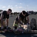 Two individuals in firefighter turnout gear (pants with reflective yellow stripes) and black shirts are attending to a person in military camouflage uniform lying on a paved surface. The scene appears to be a mass casualty or emergency response training exercise, evidenced by simulated blood/moulage on the casualty and an ambulance visible in the background. Multiple other personnel are visible in the background engaged in similar activities.