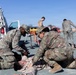 Military medical personnel in camouflage uniforms are conducting what appears to be a combat casualty care training exercise or drill. Two soldiers in the foreground are treating a simulated casualty lying on the ground, while additional personnel and equipment are visible in the background. An ambulance with its rear doors open is positioned nearby, along with a stretcher and other medical equipment.