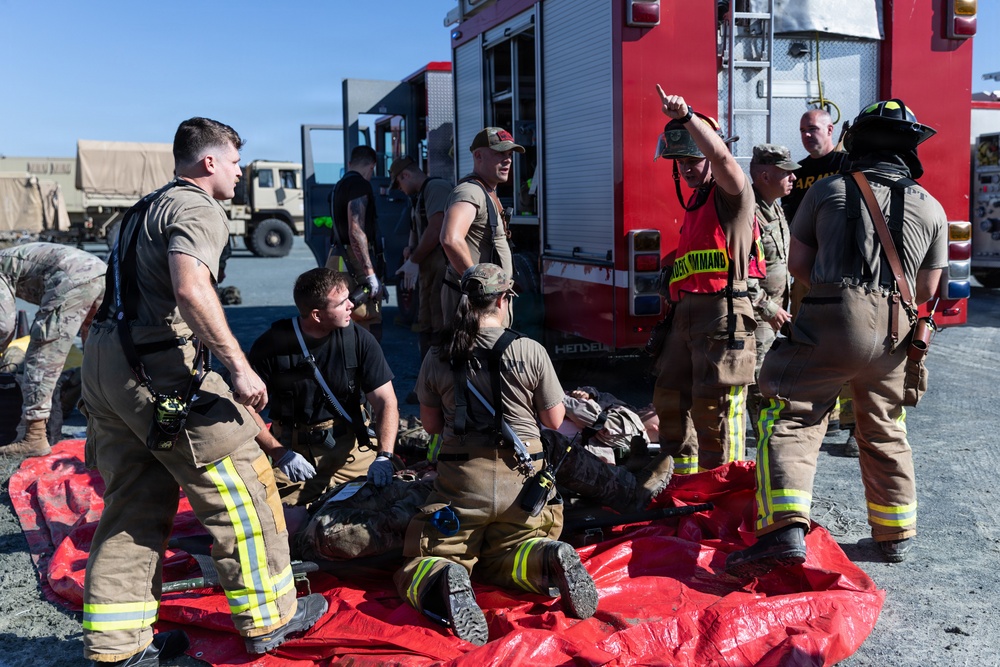 A group of military firefighters and emergency responders are conducting what appears to be a mass casualty training exercise. Personnel in firefighting gear with yellow reflective stripes are attending to simulated casualties lying on red tarpaulins on the ground. A person wearing a red 'INCIDENT COMMAND' vest is directing operations near a red fire apparatus vehicle.