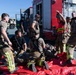 A group of military firefighters and emergency responders are conducting what appears to be a mass casualty training exercise. Personnel in firefighting gear with yellow reflective stripes are attending to simulated casualties lying on red tarpaulins on the ground. A person wearing a red 'INCIDENT COMMAND' vest is directing operations near a red fire apparatus vehicle.