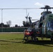 A military medical evacuation (MEDEVAC) operation is underway beside a camouflage-painted military helicopter, likely an IAR 330 Puma or similar variant. Personnel in military flight suits are loading or unloading an orange stretcher/gurney near the helicopter's side door. The scene takes place on a grass field adjacent to what appears to be a military airbase or installation.