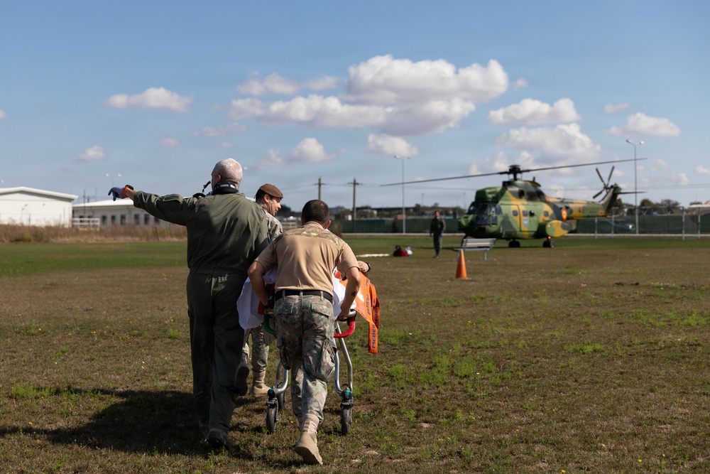 Military or emergency personnel are transporting a patient on a wheeled stretcher across a grass field toward a military helicopter. The helicopter, painted in green and yellow camouflage, appears to be on standby for a medical evacuation (MEDEVAC) operation. At least three personnel are involved in moving the stretcher, with one individual pointing toward the aircraft.