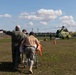 Military or emergency personnel are transporting a patient on a wheeled stretcher across a grass field toward a military helicopter. The helicopter, painted in green and yellow camouflage, appears to be on standby for a medical evacuation (MEDEVAC) operation. At least three personnel are involved in moving the stretcher, with one individual pointing toward the aircraft.