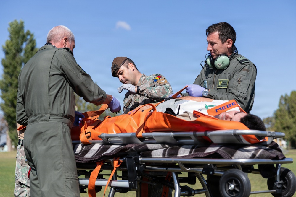 Three military personnel in green flight suits and combat uniforms are attending to a patient on an orange stretcher outdoors. The scene appears to depict a medical evacuation or emergency response exercise, with personnel wearing gloves and aviation headsets. The setting is a grassy outdoor area under clear blue skies.